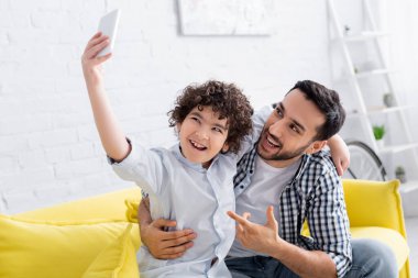 Heyecanlı Arap adam mutlu oğlunu işaret ederek akıllı telefondan selfie çekiyor.