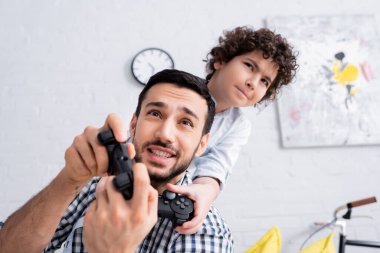 KYIV, UKRAINE - JANUARY 15, 2021: excited muslim father and son playing video game at home clipart