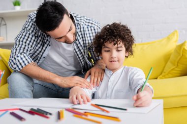 young arabian man touching son drawing with pencil in sketchbook on blurred foreground clipart