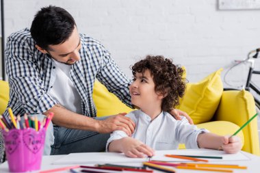 muslim man touching shoulders of cheerful son holding pencil on blurred foreground clipart