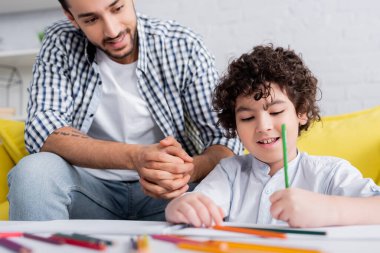 smiling muslim man sitting near son drawing with pencil on blurred foreground clipart