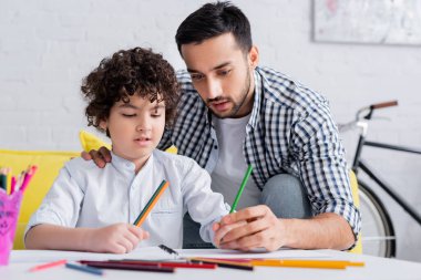 Genç Müslüman adam oğlunun bulanık ön planda kalemle çizmesine yardım ediyor.