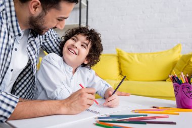 joyful muslim kid and father looking at each other while drawing with pencils together clipart