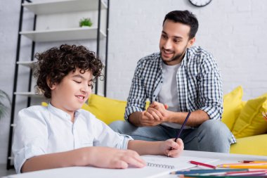 happy arabian man looking at smiling child drawing with pencil at home clipart