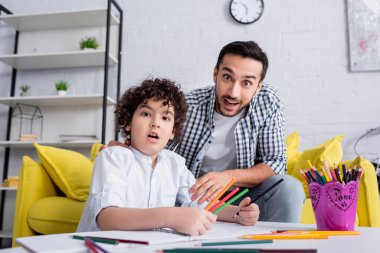 amazed muslim boy holding color pencils and looking at camera near excited father, blurred foreground clipart