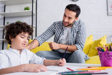 happy arabian man sitting near smiling son drawing with pencil on blurred foreground clipart