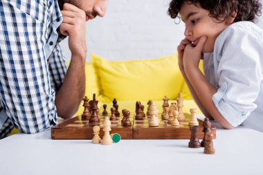 Chess on board near pensive arabian boy playing with father on blurred background  clipart