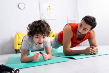 Dambılların yanında duran gülümseyen Arap çocuk ve bulanık arka planda dilini çıkaran baba. 