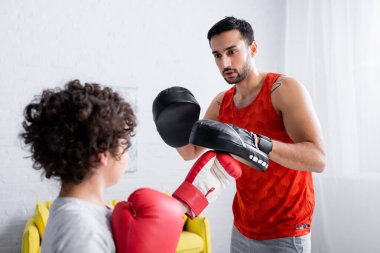 Arabian man in punch mitts training with son in boxing gloves on blurred foreground  clipart