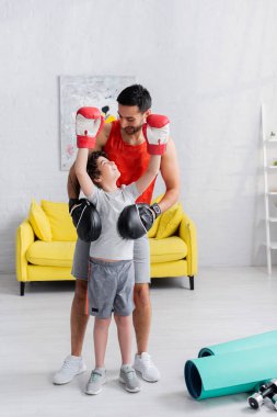 Muslim boy in boxing gloves looking at father near fitness mats and dumbbells  clipart