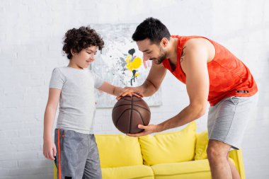 Smiling arabian man with basketball teaching son at home  clipart