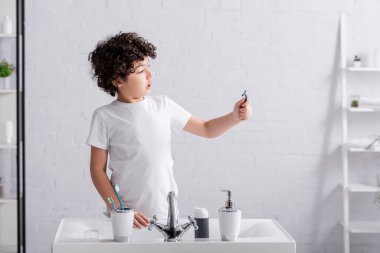 Excited muslim boy with shaving foam on nose looking at razor in bathroom  clipart