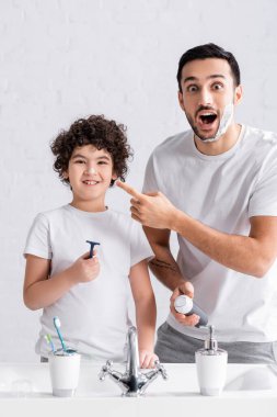 Excited muslim man holding shaving foam near smiling son with razor in bathroom  clipart