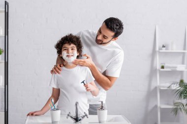 Gülümseyen Arap baba banyoda tıraş köpüğü ve tıraş köpüğü kullanan çocuk. 