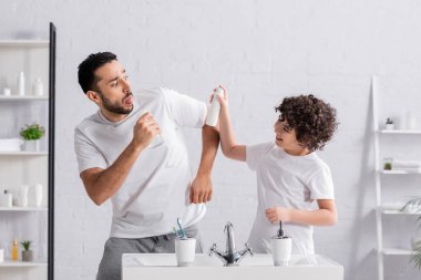 Gülümseyen Arap çocuk babasının yanında deodorant tutuyor ve lavabo açıyor. 