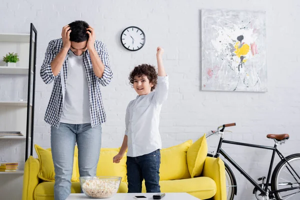 tired arabian man touching head while standing near cheerful son showing win gesture