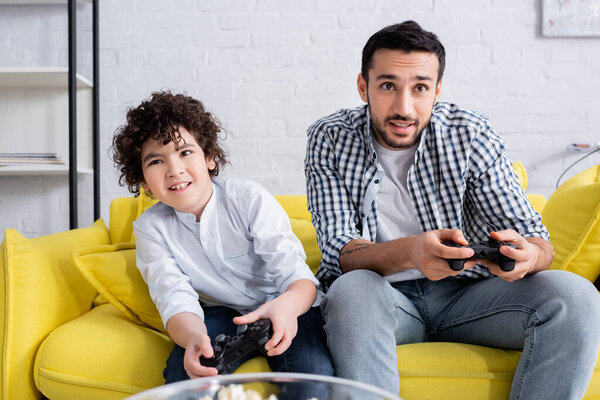 КИЕВ, УКРАИНА - 15 ЯНВАРЯ 2021: сконцентрированные мусульманский папа и сын игры с джойстиками дома