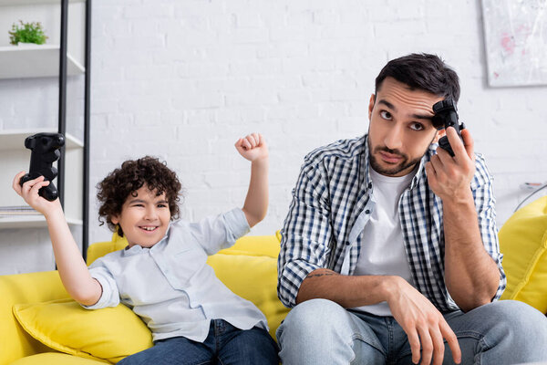 КИЕВ, УКРАИНА - 15 ЯНВАРЯ 2021: взволнованный арабский ребенок держит джойстик и показывает победный жест рядом с оскорбленным отцом