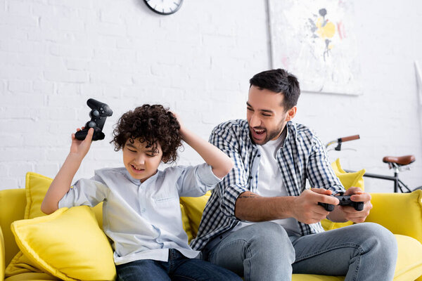 КИЕВ, УКРАИНА - 15 ЯНВАРЯ 2021: Мусульманин смеется над унылым сыном, касающимся головы, держа в руках джойстик