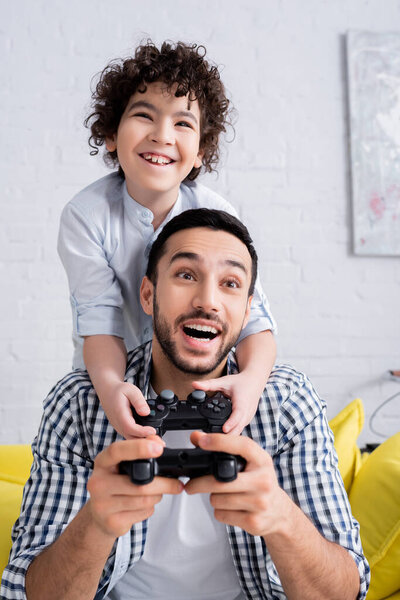 КИЕВ, УКРАИНА - 15 ЯНВАРЯ 2021: изумленный арабский мужчина, играющий в видеоигры вместе с взволнованным сыном