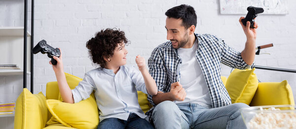 КИЕВ, УКРАИНА - 15 ЯНВАРЯ 2021 года: взволнованные арабский отец и сын, держащие в руках джойстики и показывающие победный жест, баннер