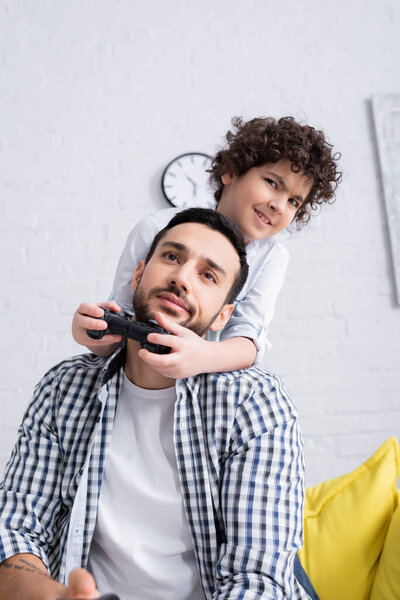 КИЕВ, УКРАИНА - 15 ЯНВАРЯ 2021: улыбающийся мальчик-мусульманин, играющий в видеоигры рядом со скептически настроенным отцом