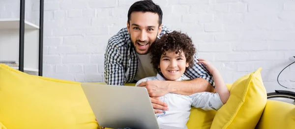 Happy Muslim Man Hugging Smiling Son Sitting Sofa Laptop Banner — Stock Photo, Image