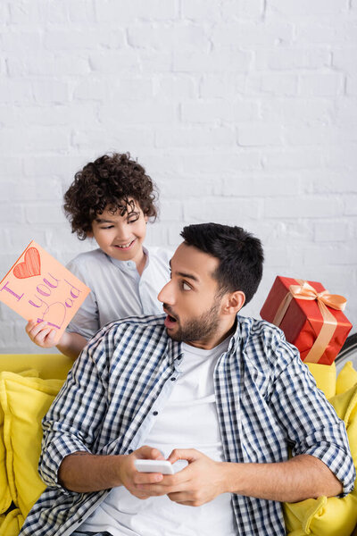 surprised muslim man near cheerful son holding i love you dad card and gift box