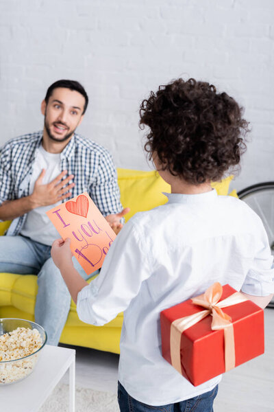 back view of boy with gift and i love you dad card near amazed father on blurred background