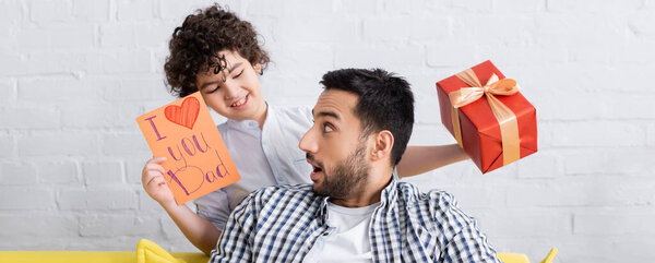 happy arabian boy holding gift box and i love you dad card near amazed father, banner