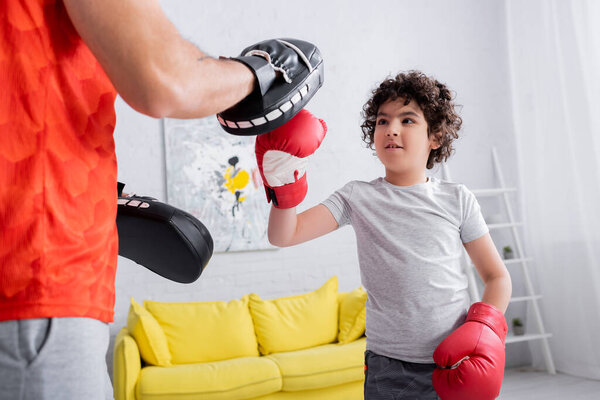 Father in punch mitts standing near muslim son in boxing gloves at home 