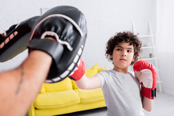 Arabian boy boxing with father in punch mitts on blurred foreground at home 