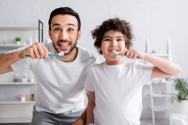 Smiling muslim father and son holding toothbrushes with toothpaste in bathroom 