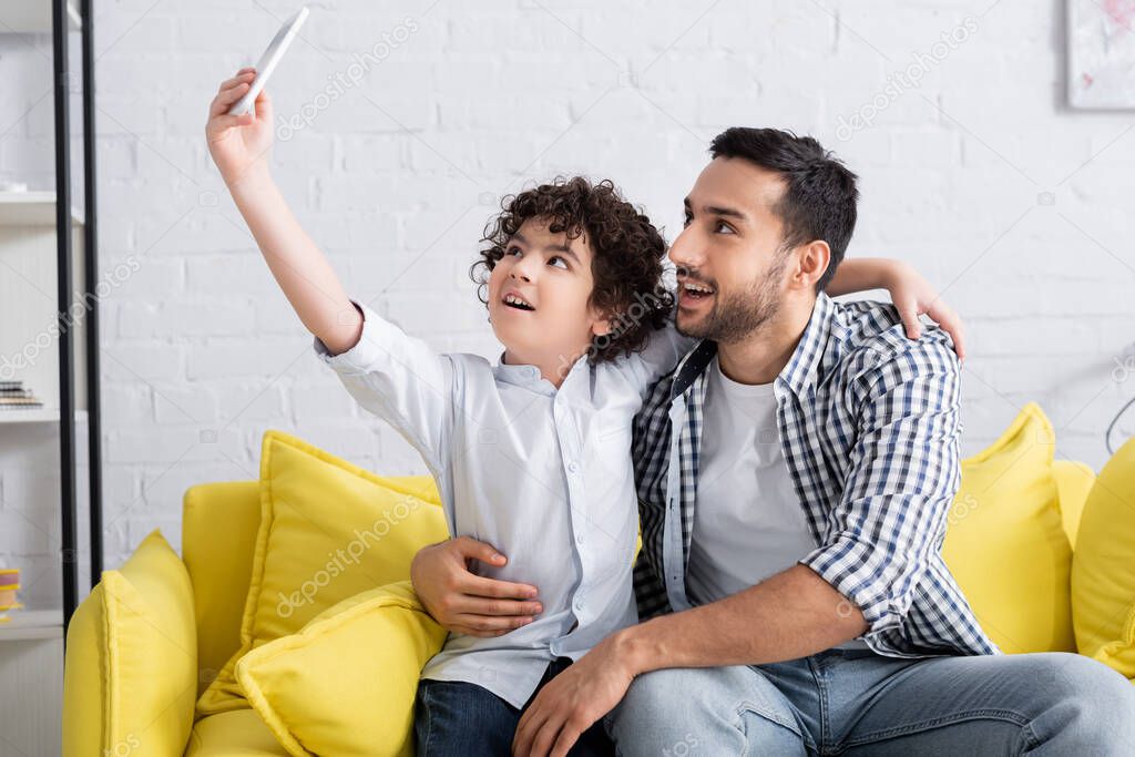 happy muslim father and son taking selfie on sofa at home