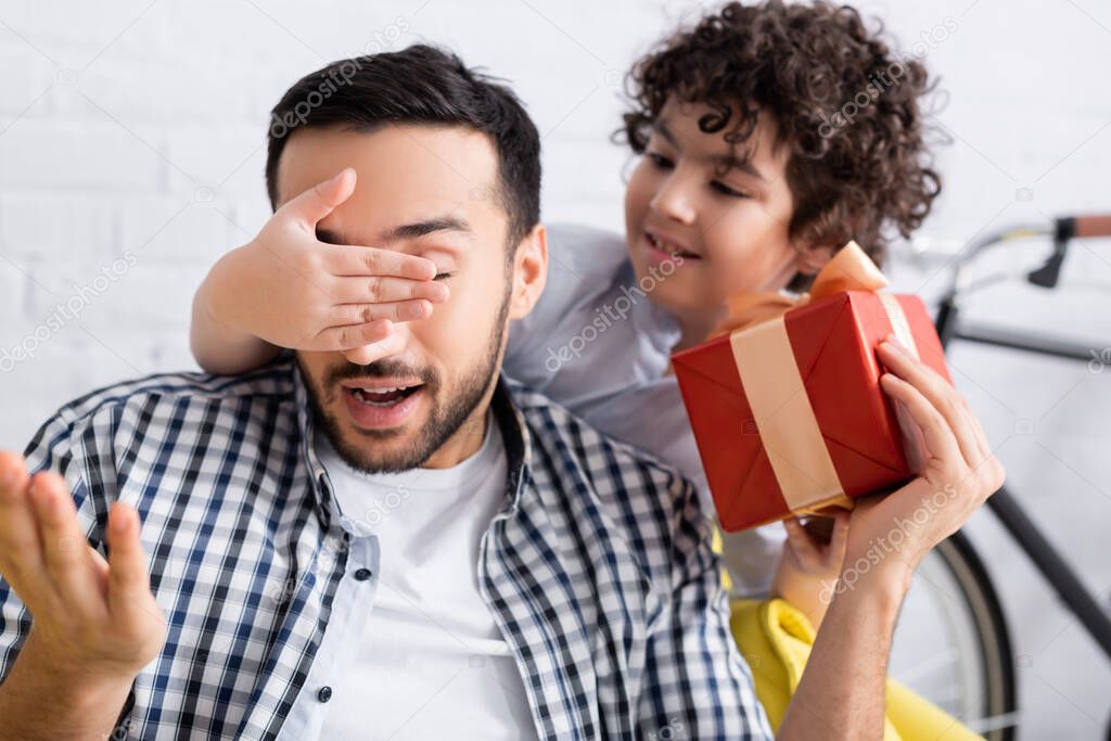 muslim boy covering eyes of father holding gift box, blurred background