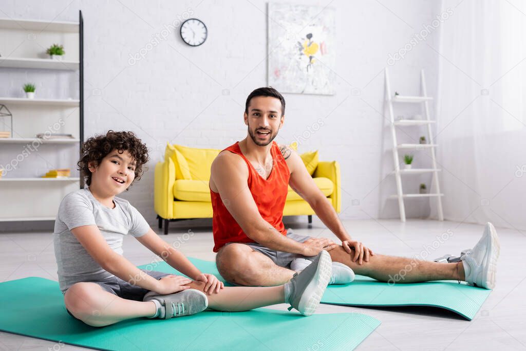 Smiling muslim son and father looking at camera while training on fitness mats 