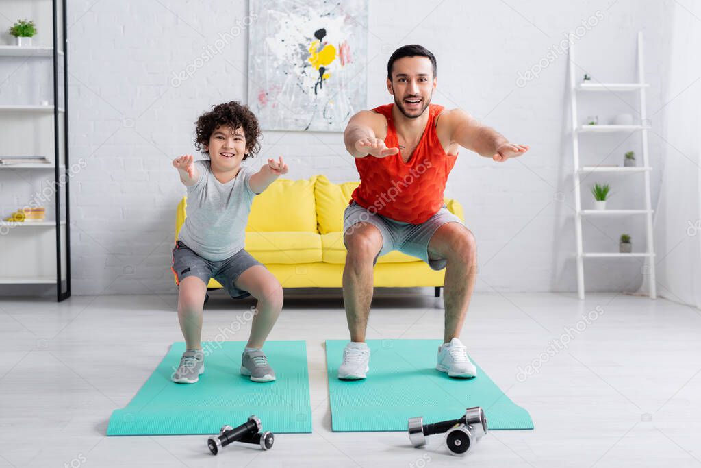 Smiling arabian son and father doing squats on fitness mats near dumbbells 