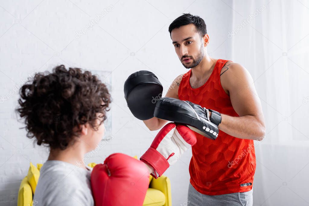 Arabian man in punch mitts training with son in boxing gloves on blurred foreground 