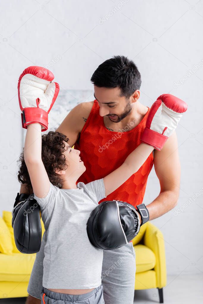 Arabian man standing near cheerful son in boxing gloves at home 