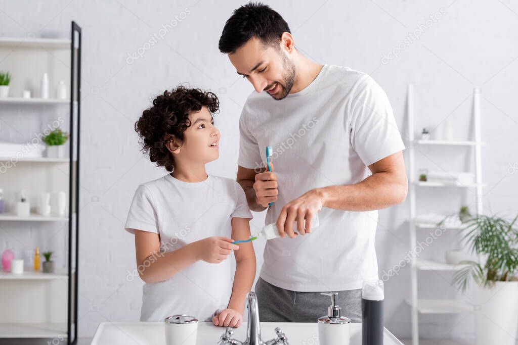 Smiling father applying toothpaste on toothbrush near muslim son and sink 