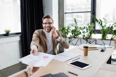 Gülümseyen işadamı akıllı telefondan konuşuyor ve bulanık arkaplanda meslektaşından evrakları alıyor. 