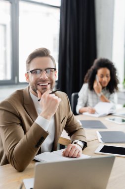 Not defteri ve dizüstü bilgisayarının yanında kameraya gülümseyen kalemli genç bir iş adamı. 