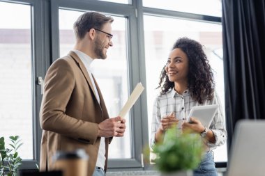 Ofiste konuşan dijital tabletli ve kağıt klasörlü neşeli çok ırklı iş adamları 