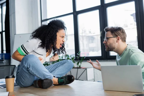 Angry interracial business people quarrelling near coffee to go and laptop