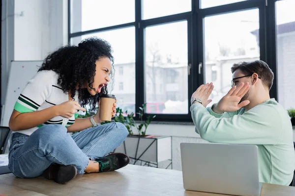 Businessman showing stop gesture to angry african american colleague with coffee to go in office