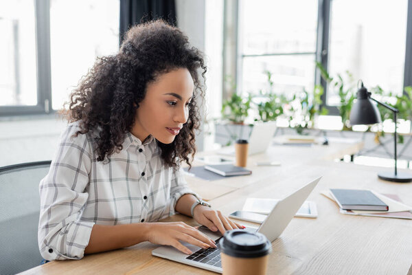 Африканская американская деловая женщина использует ноутбук рядом с кофе, чтобы выйти на размытый передний план 