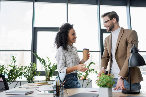 Улыбающийся бизнесмен смотрит на африканского американского коллегу с напитком на вынос у стола в офисе 