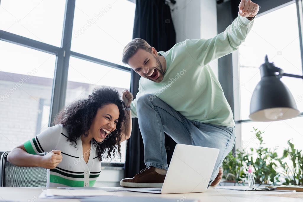 Excited interracial colleagues looking at laptop in office 