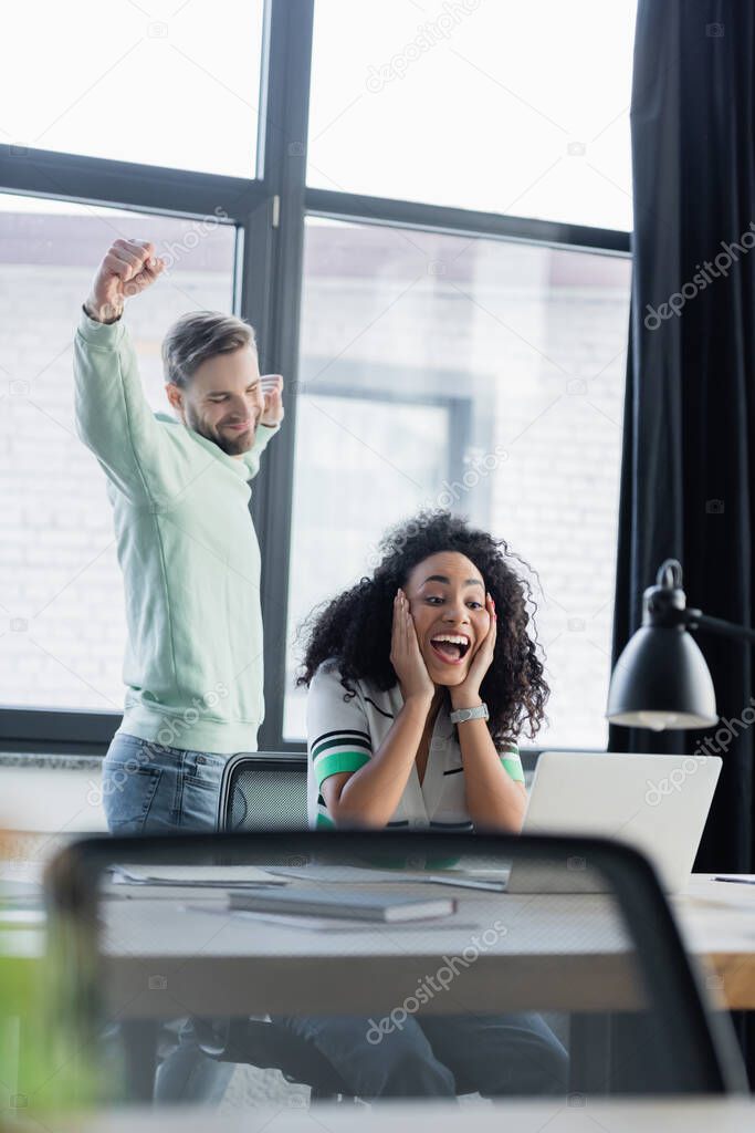 Excited african american woman looking at laptop near businessman showing yes gesture in office 