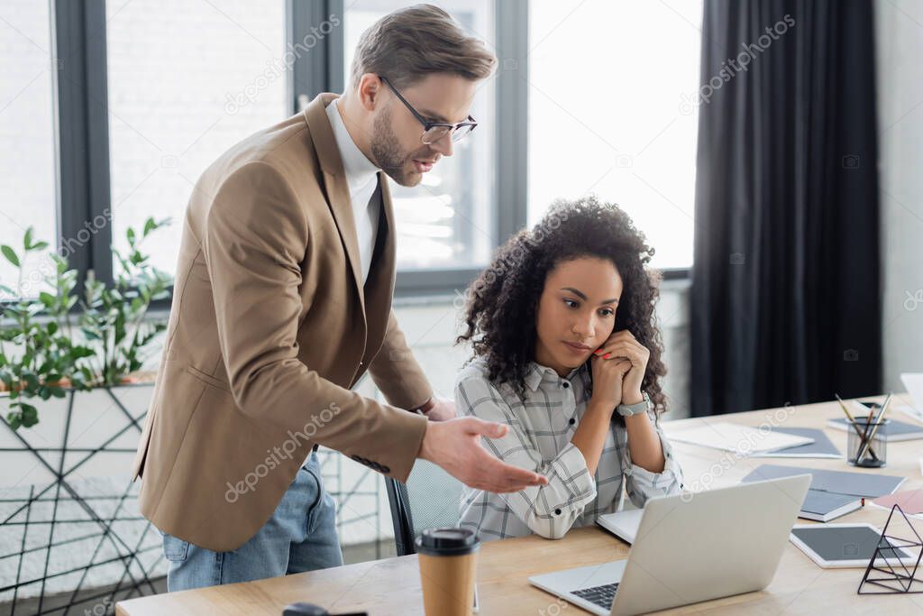 Businessman pointing at laptop near african american colleague in office 